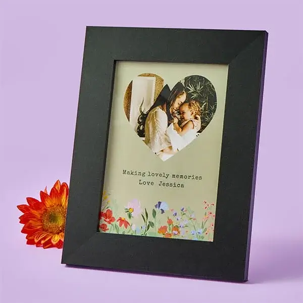 A black photo frame displaying a heart‑shaped photo cutout of a woman sharing a warm moment with a child, with a floral design and a personalised message at the bottom, shown beside an orange flower.